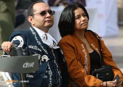 ReelAbilities Los Angeles 2022. A man and a seated outside facing forward. The man is wearing a traditional Mariachi suit and holding a roller walker. The woman is wearing a light brown long-sleeve shirt with white embroidery. Both have slight smiles on their faces.